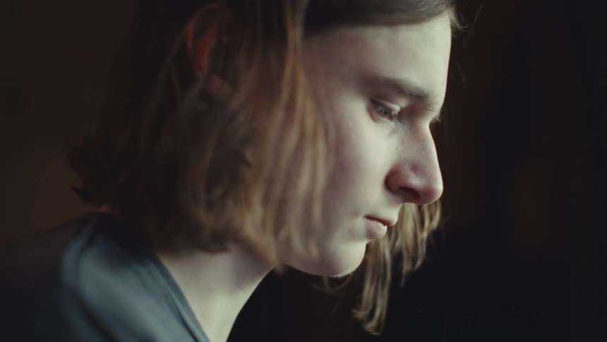 A pensive young person with shoulder-length hair is shown in a moody close-up, deeply focused on something off-screen