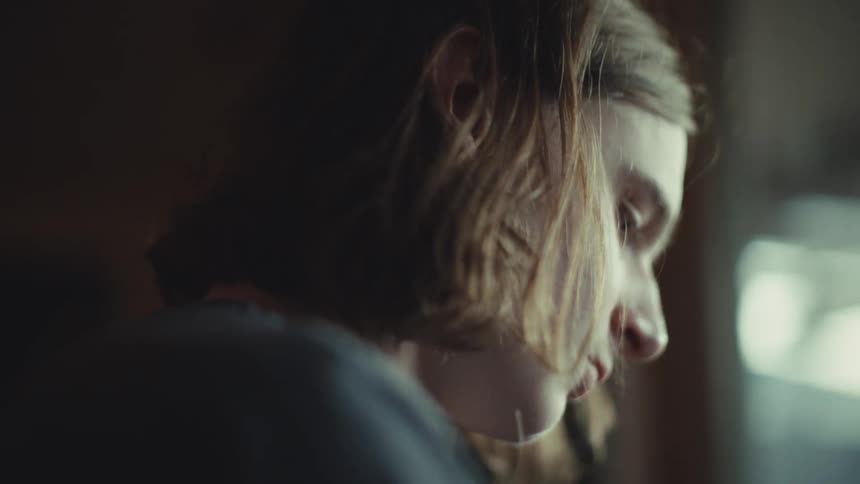 A pensive young person with shoulder-length hair is shown in a moody close-up, deeply focused on something off-screen