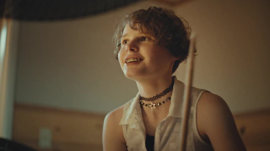 A happy young drummer with short curly hair and braces passionately plays the drums in a warmly lit room, shown in a low-angle shot