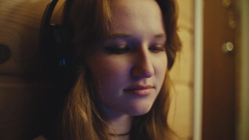 Close up portrait of a calm young woman peacefully listening to music on her headphones in a dimly lit room