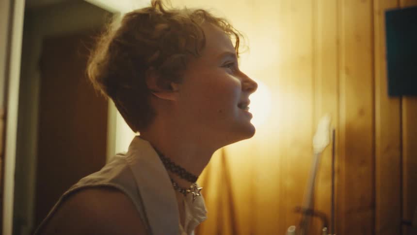 A joyful young musician with a punk aesthetic smiles while playing the drums in a warmly lit home studio