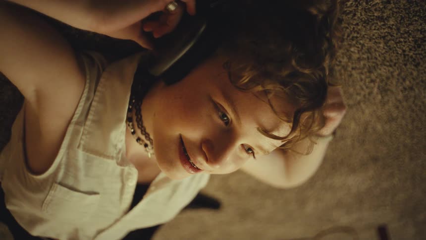 Overhead shot of a smiling teenager with braces and curly hair listening to music on headphones while lying on the floor
