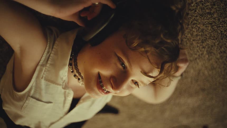Overhead shot of a smiling teenager with braces and curly hair listening to music on headphones while lying on the floor