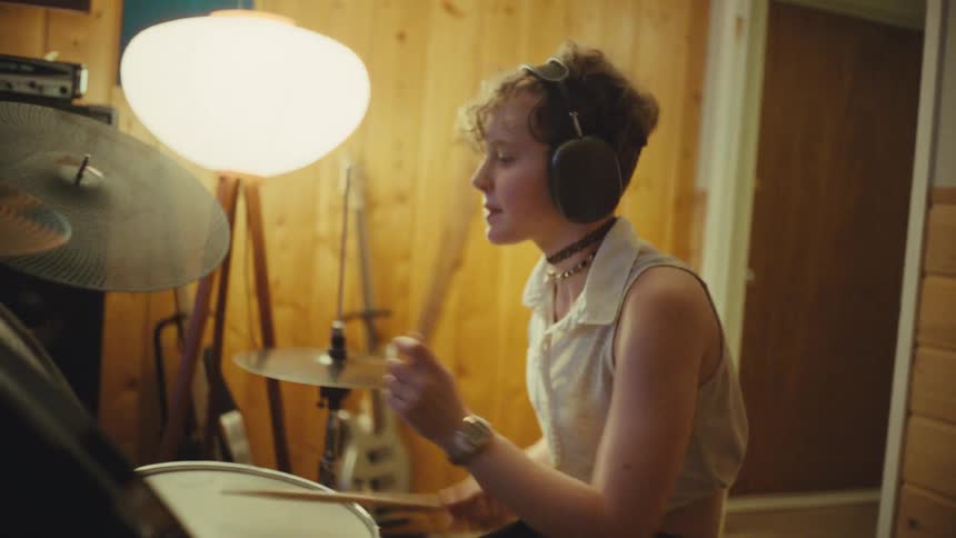 A young musician with headphones energetically plays a drum kit in a cozy, wood-paneled room