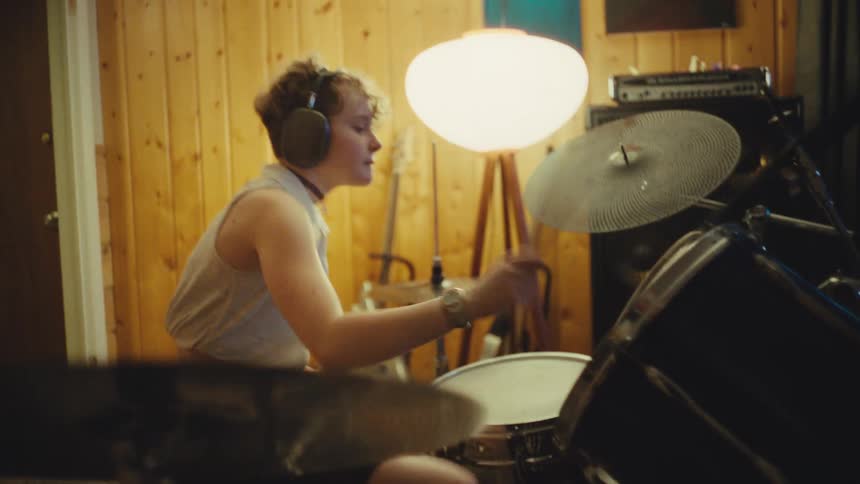 A talented young drummer wearing headphones energetically plays a beat on their drum kit in a cozy, wood-paneled music studio