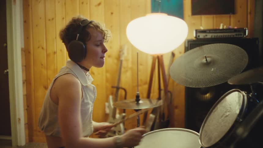 A talented young drummer wearing headphones energetically plays a beat on their drum kit in a cozy, wood-paneled music studio