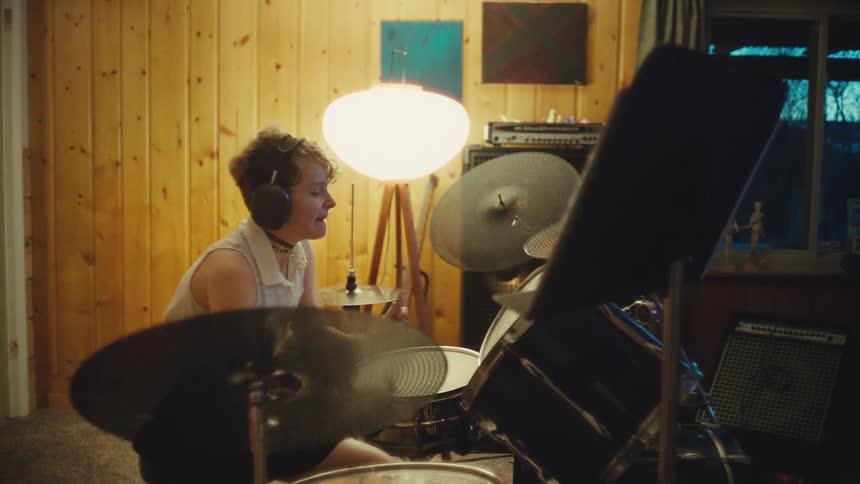 A stylish young musician with headphones energetically plays the drums in a cozy, wood-paneled home studio