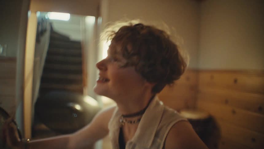 A smiling teenager with braces and short hair joyfully plays the drums in a sunlit basement practice room