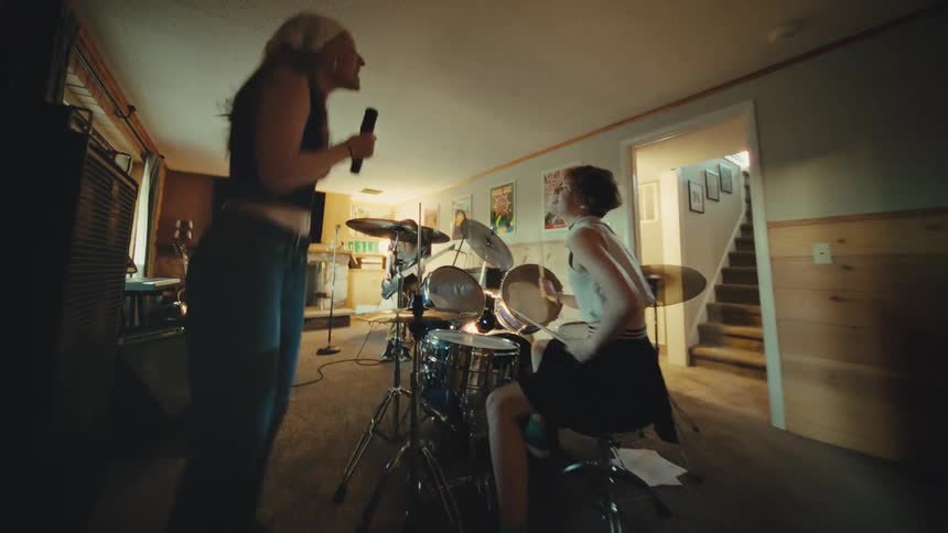 A young and energetic rock band practices their music in a home studio, with the singer dancing and the drummer playing intensely