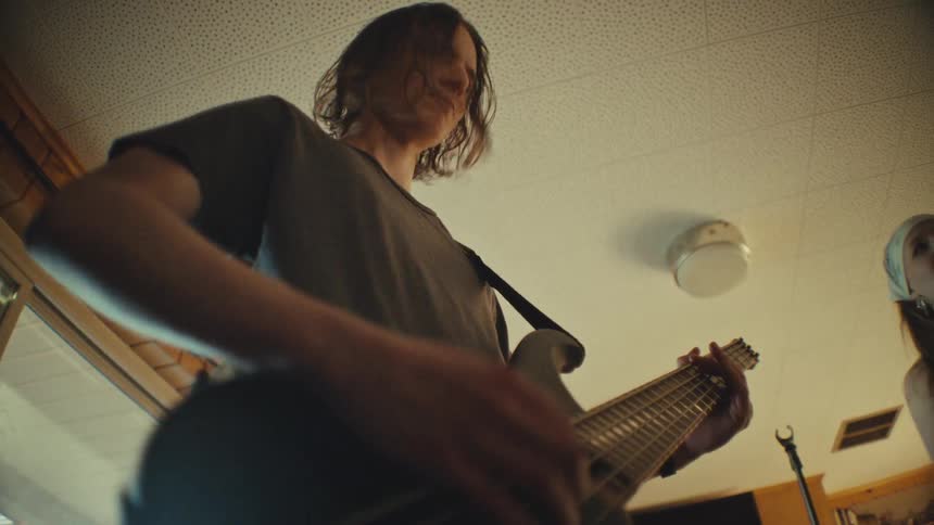 Energetic low-angle slow-motion shot of a bassist playing a five-string guitar while the lead singer passionately performs in a home studio