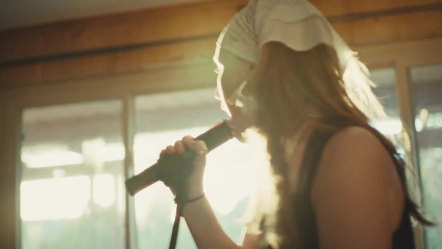 A passionate young woman in a bandana sings into a microphone, her silhouette backlit by the bright, hazy sun creating a beautiful lens flare