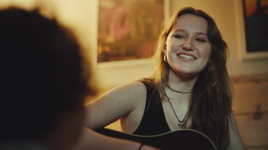 Joyful young female musician sings and plays an acoustic guitar for a friend in a warmly lit, cozy room