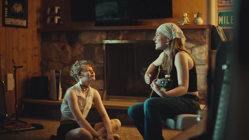 A young woman plays a song on her acoustic guitar for her friend as they sing together in a cozy, rustic room