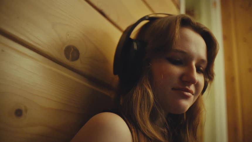 A serene young woman with headphones on leans against a wooden wall, lost in the music she's listening to