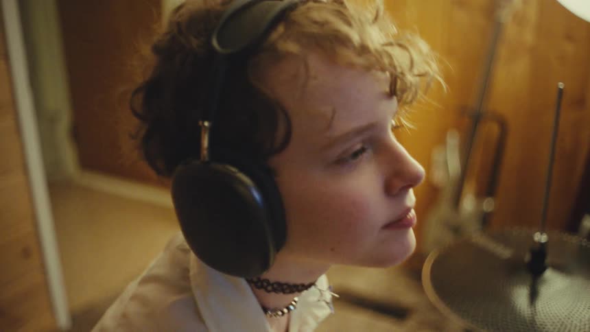 A cool young drummer with curly hair wears headphones and practices on their drum set in a cozy, warm-lit room