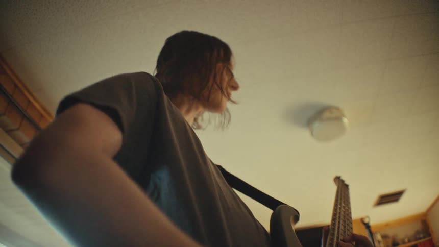Low-angle slow-motion shot of a bassist playing an electric bass guitar during a band rehearsal in a sunlit room