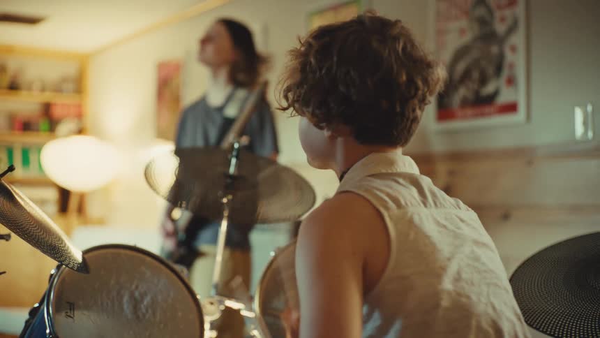 A young drummer with curly hair plays energetically during a band practice session with a guitarist in a cozy, poster-filled room