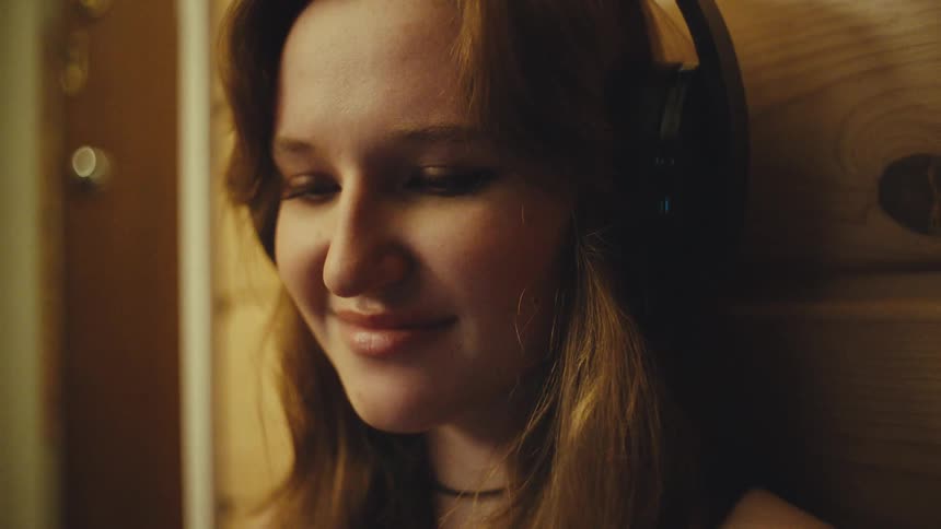 A happy young woman listens to music on her headphones in a dimly lit room and smiles