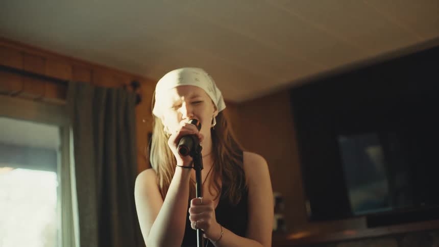 A female singer wearing a white bandana passionately sings into a microphone in slow motion while headbanging in a sunlit room