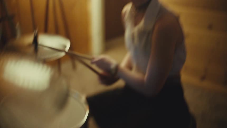 A young female drummer energetically plays a beat on her drum kit in a warmly lit, wood-paneled room