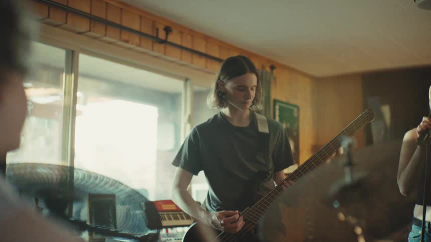 An indie rock band jams together in their home rehearsal space, seen from behind the drummer as the bassist and singer come into focus