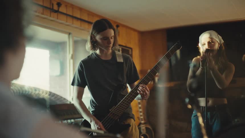 An indie rock band jams together in their home rehearsal space, seen from behind the drummer as the bassist and singer come into focus