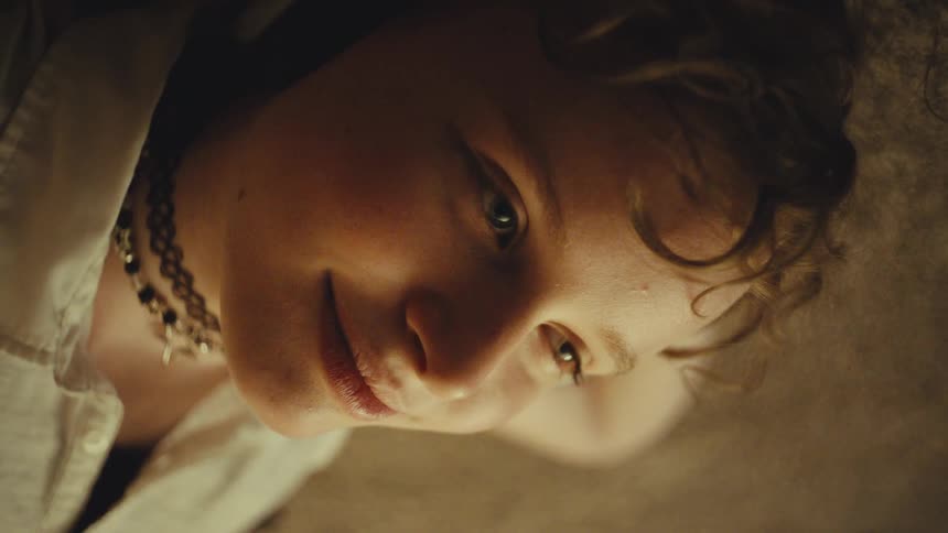 A young person with curly hair smiles gently while looking down, seen in a warm, intimate low-angle close-up
