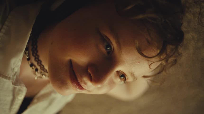 A young person with curly hair smiles gently while looking down, seen in a warm, intimate low-angle close-up