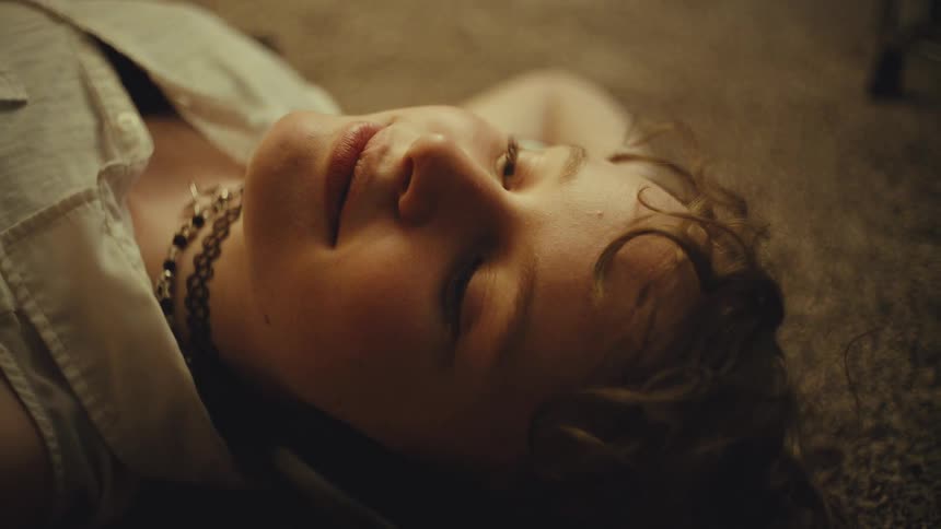 A thoughtful young woman with curly hair closes her eyes peacefully while lying on a carpeted floor in a warmly lit room