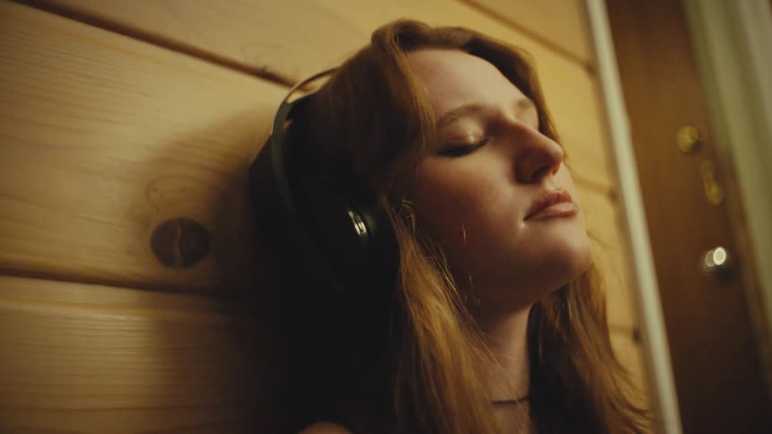 Close-up of a serene young woman with eyes closed, relaxing against a wooden wall while listening to music through headphones in a warmly lit room