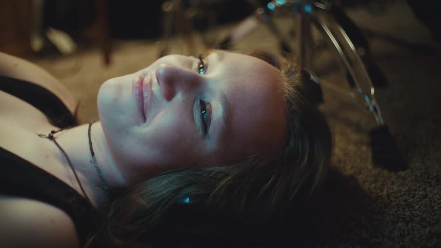 A close-up of a young woman lying on the floor in a dimly lit room as she smiles with contentment