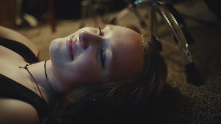 A close-up of a young woman lying on the floor in a dimly lit room as she smiles with contentment