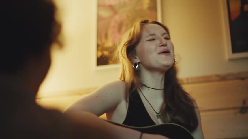 A young woman playing an acoustic guitar laughs and sings with a friend in a cozy, warmly lit room in slow motion