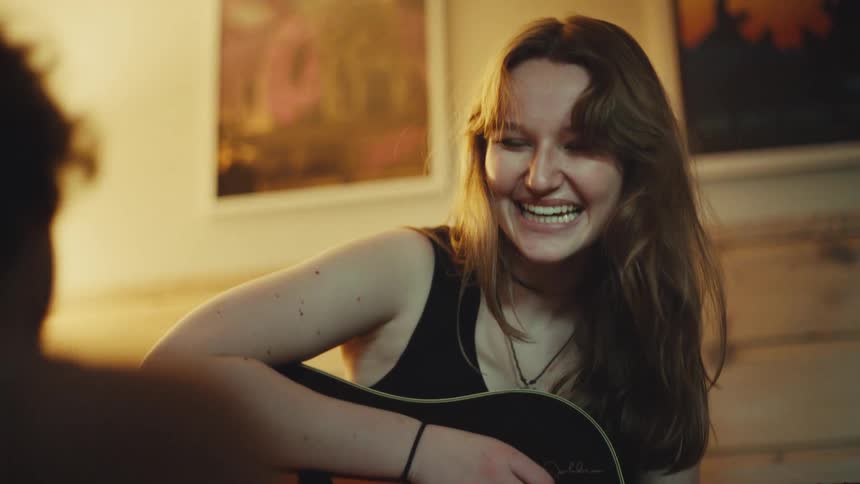 A young woman playing an acoustic guitar laughs and sings with a friend in a cozy, warmly lit room in slow motion