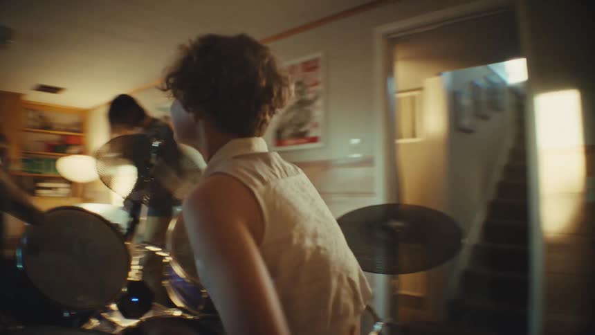 A dynamic tracking shot follows a young, energetic drummer as they play with their band in a warmly lit, wood-paneled room