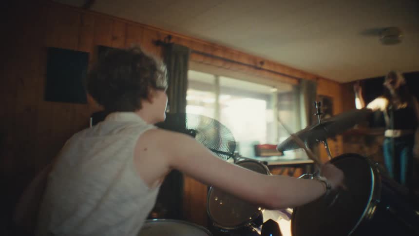 A dynamic tracking shot follows a young, energetic drummer as they play with their band in a warmly lit, wood-paneled room