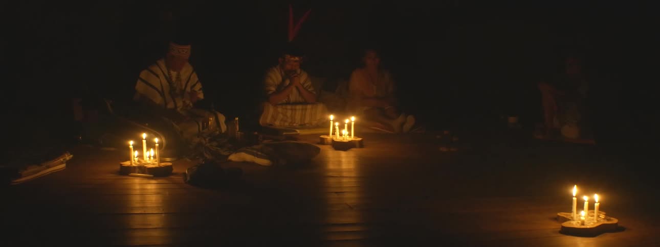 Shamans in traditional attire lead a spiritual healing ceremony in a dark room illuminated only by candles