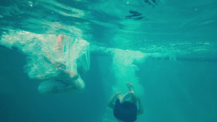 Two young boys jump one after another into a resort swimming pool in a fun slow-motion shot that goes underwater
