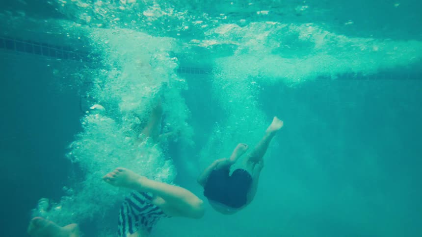 Two young boys jump one after another into a resort swimming pool in a fun slow-motion shot that goes underwater