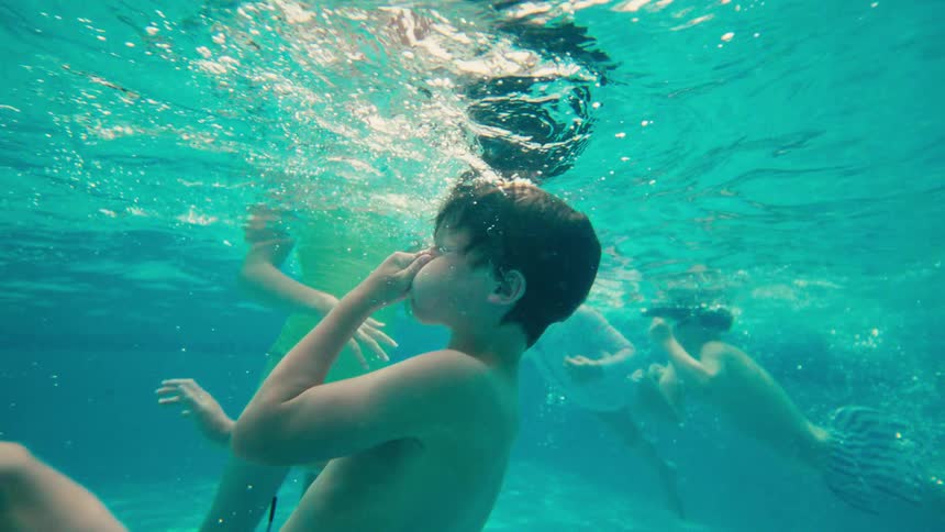 A slow-motion underwater view shows several children playing and swimming in a clear turquoise pool