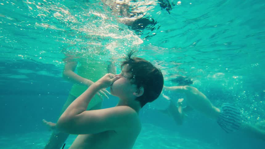 A slow-motion underwater view shows several children playing and swimming in a clear turquoise pool