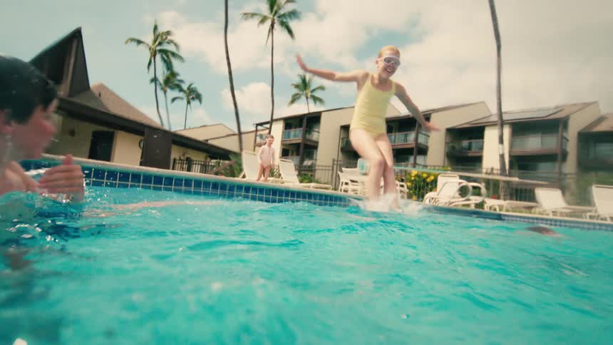 A young girl wearing goggles joyfully jumps into a pool in slow motion, with an exciting underwater view of her splash