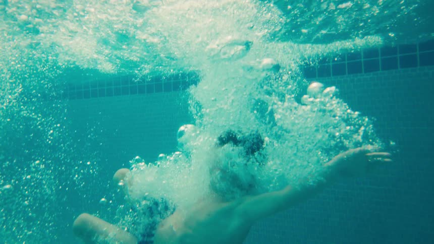 A young boy cannonballs into a swimming pool in slow motion, shot from the water's surface and then from underwater
