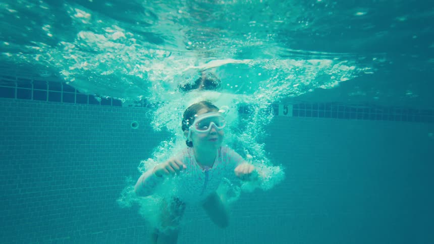 A young girl with goggles jumps into a swimming pool, filmed from an underwater perspective in slow motion