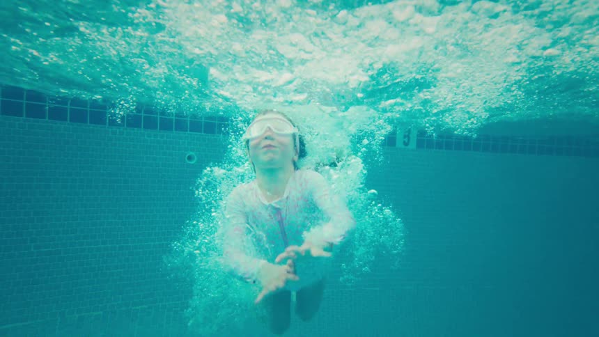 A young girl with goggles jumps into a swimming pool, filmed from an underwater perspective in slow motion