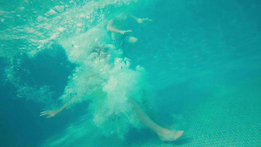 Split-level over-under shot of a person jumping into a swimming pool in super slow motion, with palm trees and a resort in the background