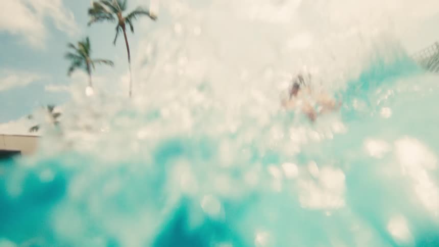 A low-angle, slow-motion shot shows a young boy doing a cannonball into a swimming pool on a sunny day