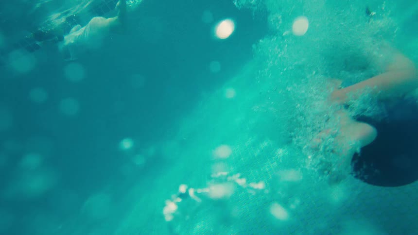 A low-angle, slow-motion shot shows a young boy doing a cannonball into a swimming pool on a sunny day