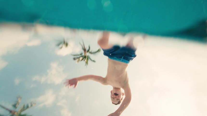 Slow motion underwater view of a happy young boy jumping with a splash into a bright blue swimming pool