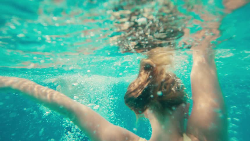 A slow-motion, underwater shot captures a woman in a swimsuit sinking into the turquoise water, her form becoming lost in the bubbles
